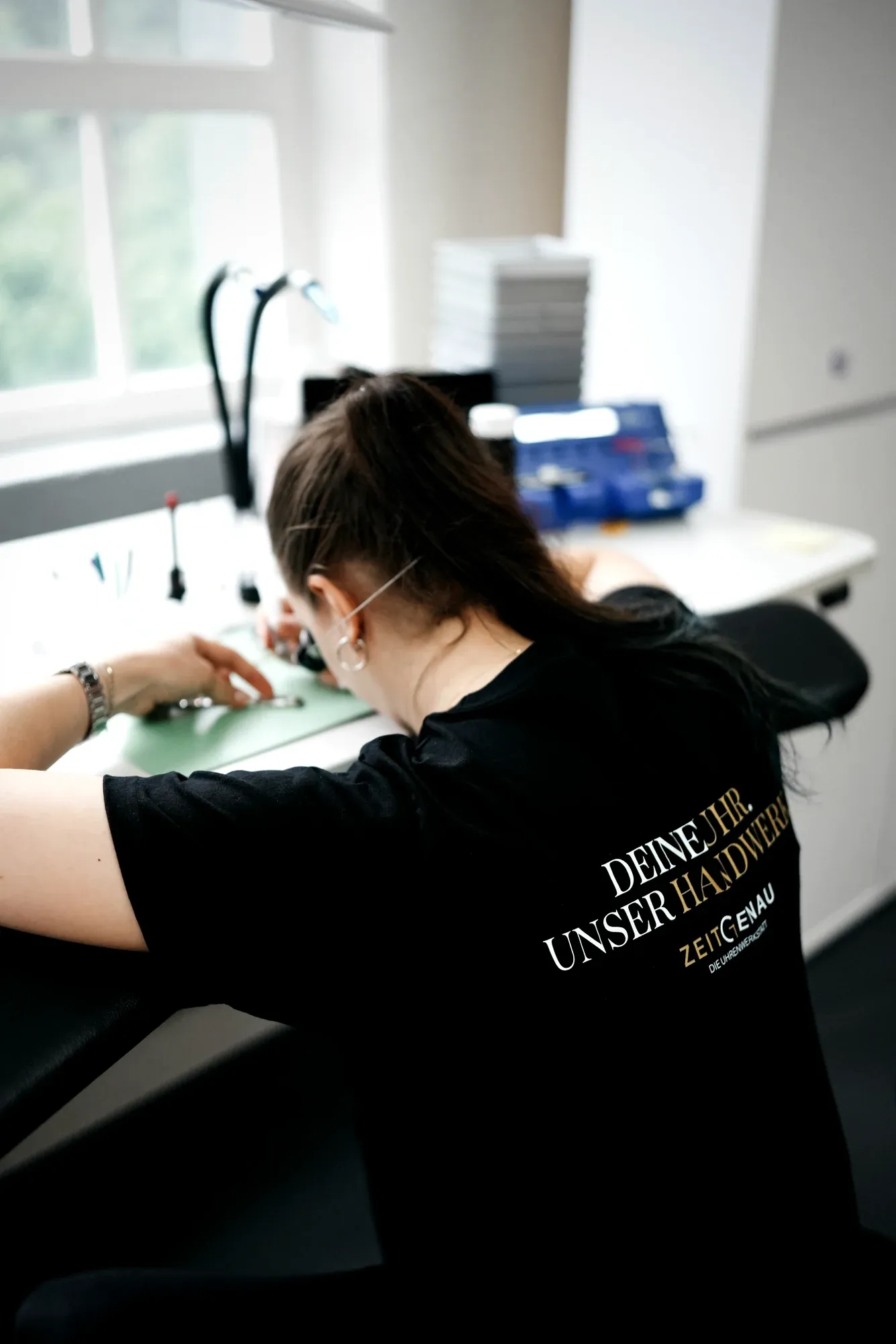Uhrmacherin am Werktisch im Zeitgenau-Shirt mit Slogan „Deine Uhr. Unser Handwerk.“ — konzentriert an der Werkzeugmatte